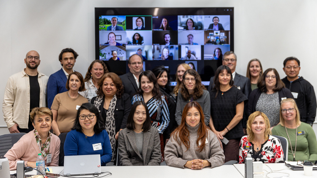 A group of about 20 researchers stand and sit for a group photo. They are in front of a tv that has remote participants shown in boxes. 