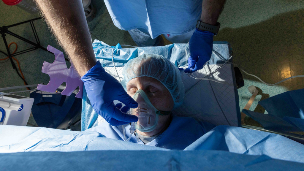 Photograph taken from above. A person with eyes closed wearing a surgical cap lies on a bed in an operating room. A gloved hand is adjusting the clear plastic mask that covers the patient's nose and mouth.