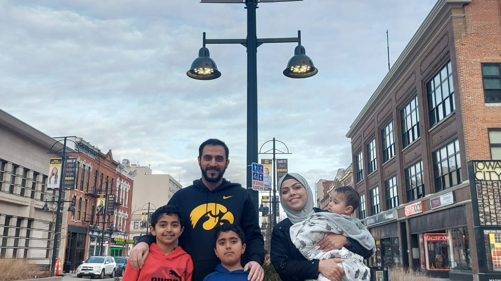 A woman holding a baby stands next to a man who has his hands on the shoulders of two boys standing in front of him. Behind the group is a pole with a banner at the top. On the banner is a photo of a woman.