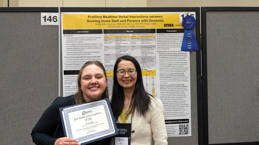 To women smile at the camera. One holds a certificate. They stand in front of a research poster with a blue ribbon.