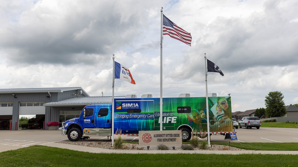 A large blue and green truck is parked behind a sign for the Alburnett Otter Creek Fire Department and three flying flags. Behind is is a low gray building. Above is a large expanse of clouds in the sky. 