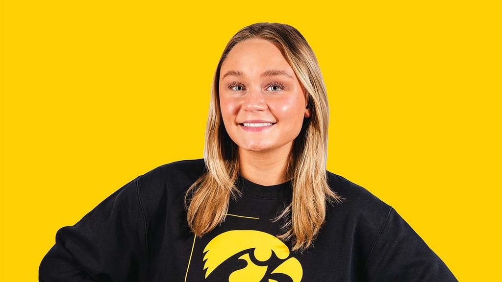 A woman wearing a black Iowa Cheer sweatshirt stands with both hands on hips,, smiling at camera. The background behind her is yellow.