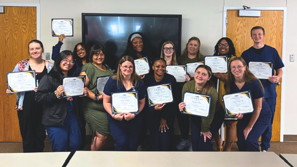 Fourteen nurse residents standing in a classroom setting, each holding a certificate of completion. Most are wearing navy blue scrubs, while a few are in casual attire. They are posed in front of a large wall-mounted screen, with wooden doors on either side and a table in the foreground.