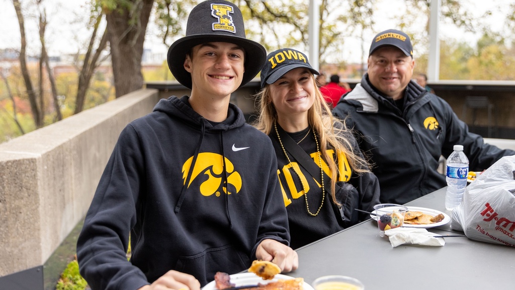 Three people in Iowa Hawkeye gear sit at a table outside. They are looking at the camera and smiling. On the table are plates of pancakes and cups of orange juice. 