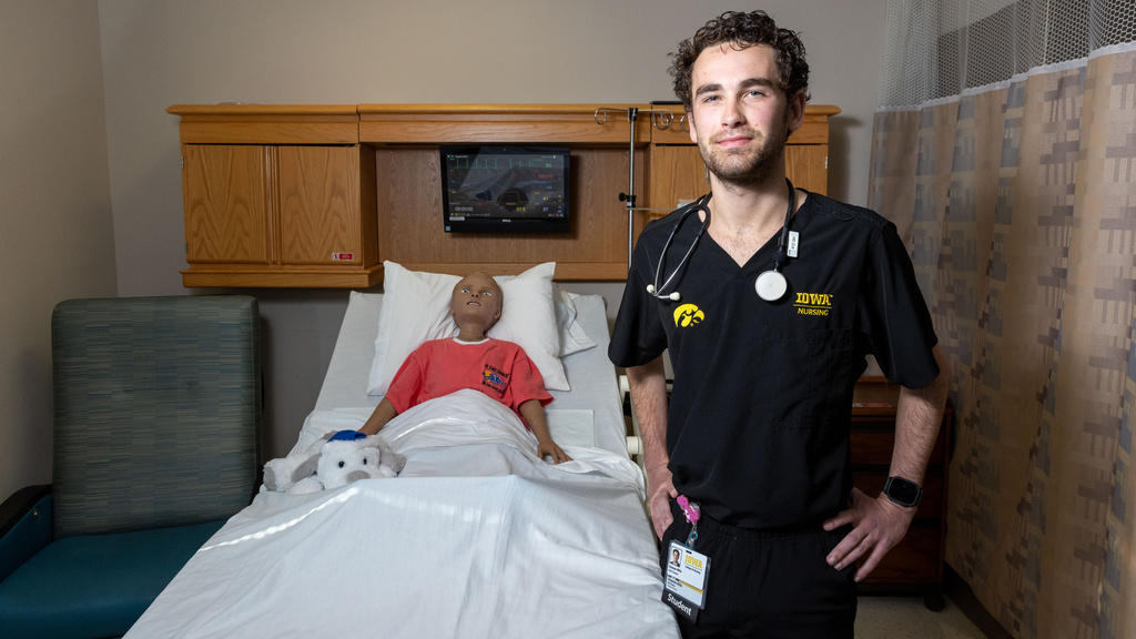 A man in black scrubs with a stethoscope around his neck poses for a portrait in a hospital room setting. On the bed next to him is a simulated patient manikin.