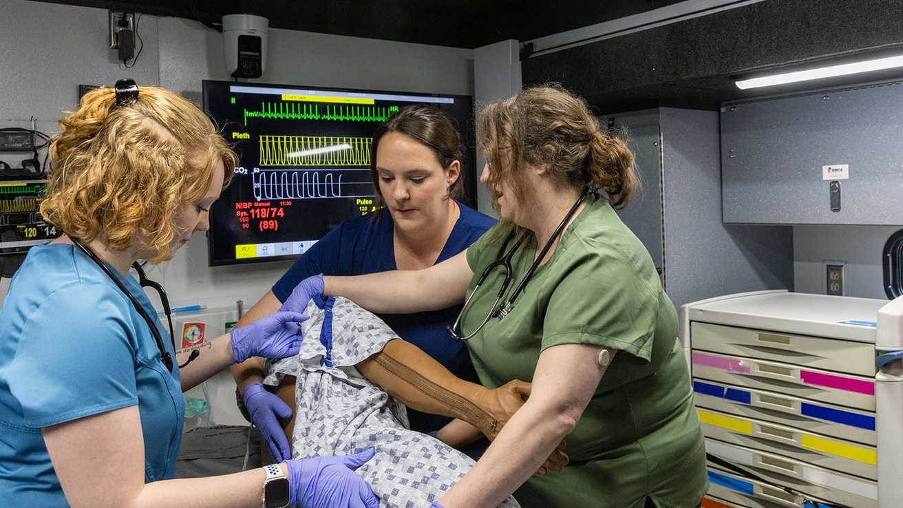 In a small area with medical equipment and a screen on the wall showing readings, three people in scrubs turn a manikin wearing a hospital gown on its side.  